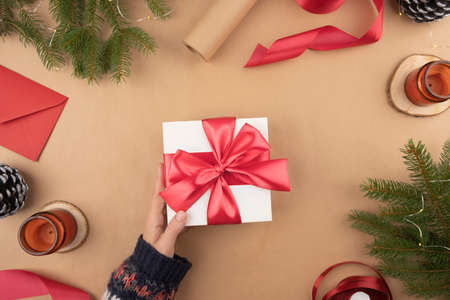 White gift box with red ribbon holds in hand on a kraft background. Packaging and preparation of gifts for the celebration. Christmas New Year backdrop with tree branch decor and lightsの写真素材