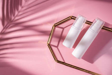 Plastic white bottles with pump for cream or lotion on glass plate for product presentation. Skin care cosmetic in top view on pink background with palm leaves shadow. Beauty concept for face careの写真素材
