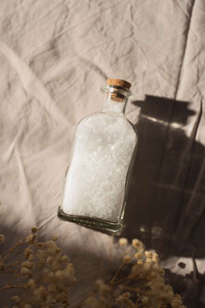 Transparent bottle filled with white bath sea salt on light grey fabric background. Beauty treatment for spa and wellness. Skincare natural cosmetic concept for body care.の写真素材