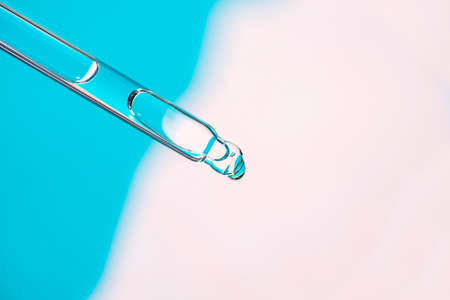 Pipette with drop of serum or cosmetic liquid close-up on blue and pink background. The appearance of the texture of the gel. Skin care products. Beauty concept for face and body careの写真素材