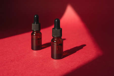 Amber bottles with dropper pipette with serum or essential oil. Red background with daylight and beautiful shadows. Skincare products, natural cosmetic. Beauty concept for face and body careの写真素材