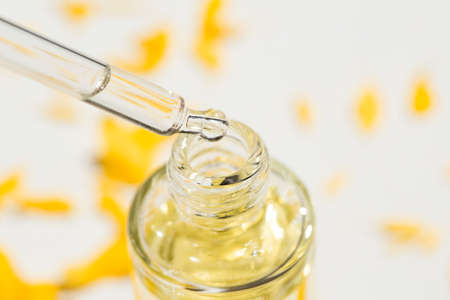 Open transparent bottle and pipette with serum or cosmetic liquid with beautiful yellow flowers in soft focus. White background with daylight and beautiful shadows. Beauty concept for face and body careの写真素材