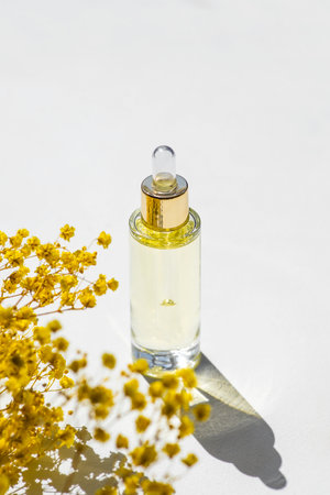 Transparent bottle with dropper pipette with serum or essential oil with beautiful yellow flowers. White background with daylight and beautiful shadows. Beauty concept for face and body careの写真素材