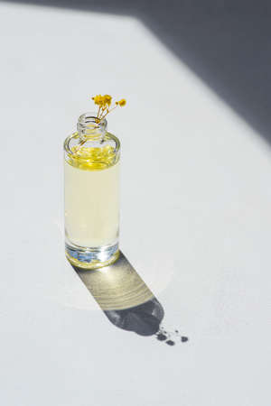 Open transparent bottle without cap with serum or essential oil with beautiful yellow flowers in soft focus. White background with daylight and beautiful shadows. Beauty concept for face and body careの写真素材