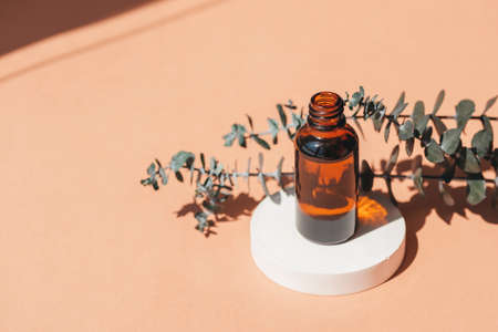 Open amber bottle without cap with eucalyptus branches. Beige background with daylight and beautiful shadows. Skincare serum or essential oil natural cosmetic. Beauty concept for face and body careの写真素材