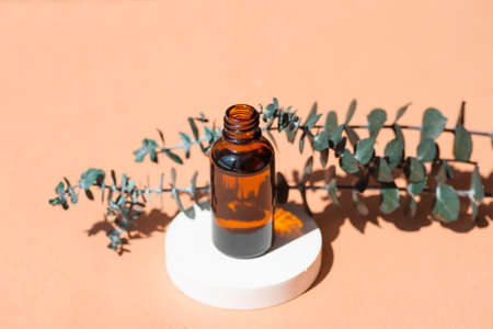 Open amber bottle without cap with eucalyptus branches. Beige background with daylight and beautiful shadows. Skincare serum or essential oil natural cosmetic. Beauty concept for face and body careの写真素材