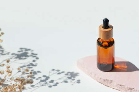 Amber bottle with cap with serum or essential oil with flowers shadows on pink platform. White background with daylight. Beauty concept for face and body care. Soothing CBD oilの写真素材