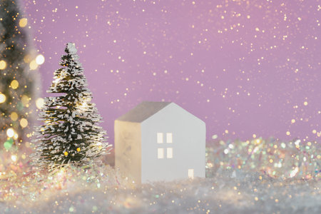 Christmas cute little house with tree in snow. Glowing festive garland on lilac background. Cozy fairytaleatmosphere with home decor with selective focus. New Year cardの写真素材