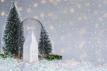 Christmas cute little house in glass dome with tree in snow. Glowing festive garland on light background. Cozy atmosphere with home decor with selective focus. New Year cardの写真素材