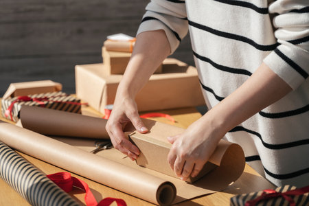 Young woman packing a box in craft paper on a wooden table. Preparation of gifts for the celebration. Giftbox wrapping in eco packaging.の写真素材
