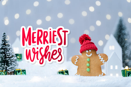 Christmas cute little house and gingerbread man with tree in snow. Cozy atmosphere with home decor with selective focus on cookie. New Year card with lettering Merriest Wishes. Glowing festive garlandの写真素材