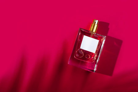 Transparent bottle of perfume with label on a red background. Fragrance presentation with daylight. Trending concept in natural materials palm leaves shadow. Womens and mens essence.の写真素材