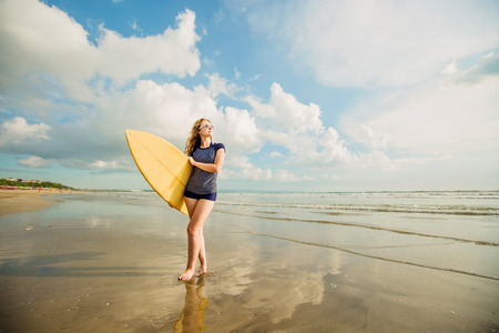 Beautiful young caucasian girl in rushwest and sunglasses with yellow surfboard at legian beach,...