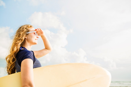 Beautiful young caucasian girl in rushwest and sunglasses with yellow surfboard at legian beach,...