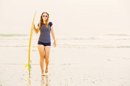 Beautiful young surfer girl stays on the beach at sunset with yellow surfboard. Healthy lifestyle, leisure, travel, holiday and sport concept, copyspaceの写真素材