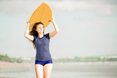 Beautiful sexy surfer girl on the beach at sunset walks along ocean shore, yellow surfboard in her...