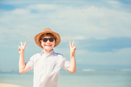 Portrait of happy red haired kid boy in hipster sunglasses and summer hat enjoying life on the beach