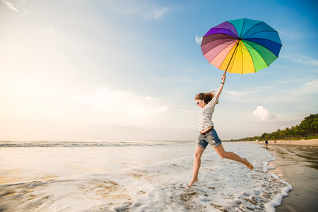 Cheerful young girl with rainbow umbrella having fun on the beach before sunset. Travel, holidays, vacation, healthy lifestyle conceptの写真素材