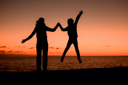 Silhouette of a young mother and her jumping son in front of a red sunset in the sky on a summer day...