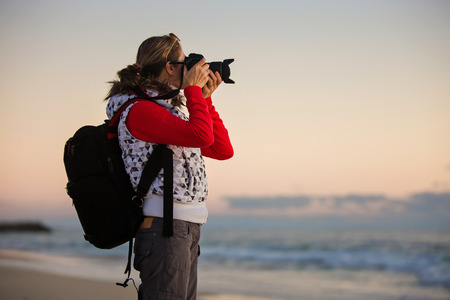 Young woman photographer with backpack taking pictures with camera at sunset on the winter beach.の写真素材