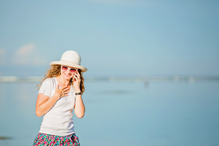 Beautiful young caucasian woman on the beach in white hat talking on mobile phone and laughing. Sunny day on summer vacationの写真素材