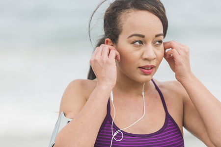 Female runner preparing for outdoor workout on the beach setting on earphones. Beautiful fit mixed race fitness model outdoorsの写真素材