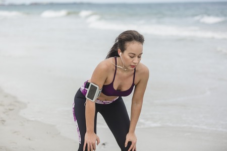 Runner woman resting after long run on the beach. Beautiful vivacious woman jogging on the beach with cloudy weatherの写真素材