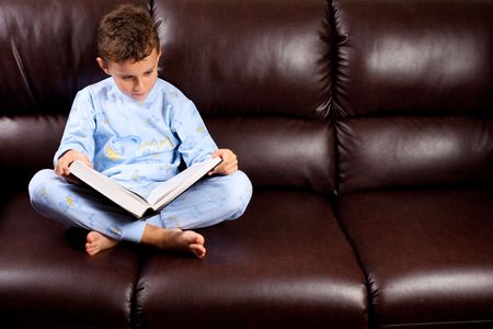 Cute schoolboy in pajamas reading a book (a dictionary) while sitting on a sofaの写真素材