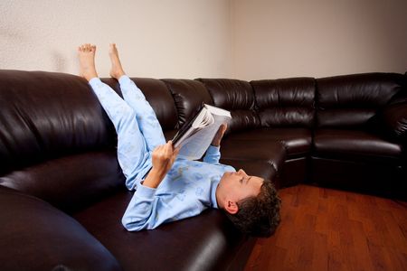 Cute schoolboy in pajamas reading a book (a dictionary) while sitting on a sofa upside downの写真素材