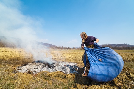 Old rural woman burning fallen leaves, spring cleaning in the garden