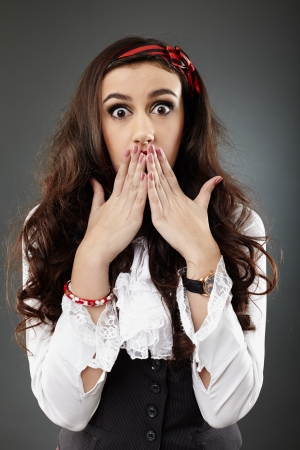 Scared young girl, covering her mouth with her hands, in closeup pose on gray backgroundの写真素材