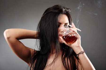 Sensual young woman drinking a glass of brandy and smoking a cigarette, closeup pose over gray background, debauchery conceptの写真素材