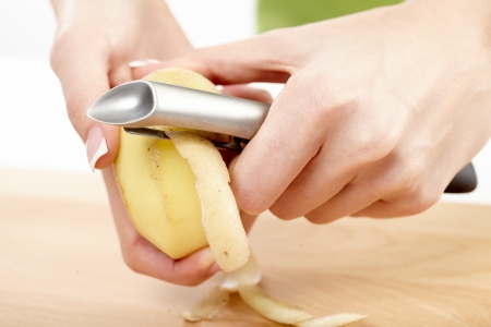 Hands of a young woman peeling potatoes with kitchen utensil on a wooden board