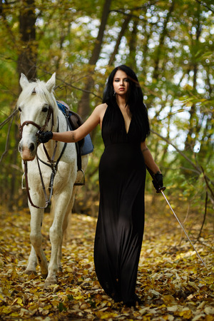 Beautiful woman with white horse and whip walking in the forest, fantasy conceptの写真素材