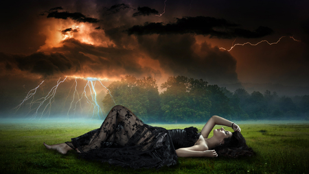 Composite image of a woman in black lace dress lying on grass with storm overheadの写真素材