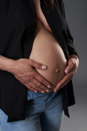 Pregnant woman in blue jeans and unbuttoned black shirt posing on gray backgroundの写真素材