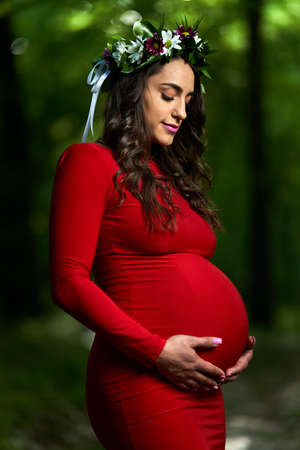 Portrait of a beautiful young hispanic pregnant woman in long red dress in a deciduous forest in the summerの写真素材