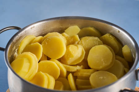 Hot freshly boiled sliced potatoes in a stainless steel pot on a blue wooden board