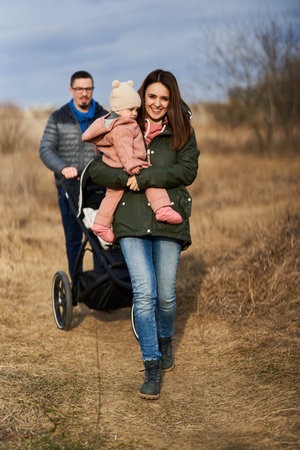 Young family with their little baby girl walking through park with baby carriage in a residential...