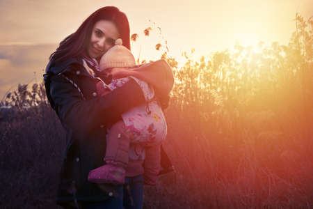 Young mother holding her sleeping baby daughter outdoor in the park in a residential area at sunsetの写真素材
