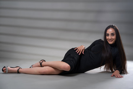 Young beautiful brunette hispanic woman with long hair in black dress posing isolated on gray...