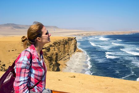 Woman looking at Ocean waves crashing into Cliff edge. Paracas National Reserve, Peru. Balletas Islands and rocks with seashore coastline. Orange, blue and green. Dramatic travel concept.の写真素材