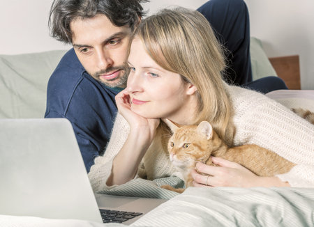 Young couple relaxing on bed with their cat using laptop to video call their family. Soft focus laptop. Love, happiness, technology, people and funの写真素材