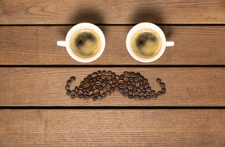 Two cup coffee and coffee beans in the shape of a mustache against a wooden background. Space for text.の写真素材
