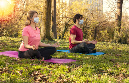 Young couple in gymsuit with Mask and Closed Eyes Sitting on Yoga Mat in Relaxing Meditation Pose at Park with Sun Reflections.の写真素材