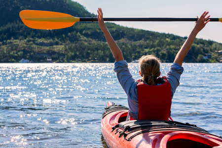 the girl holds a paddle over her head and floats along the fjord in norway, kayaking in the sea, outdoor activitiesの写真素材