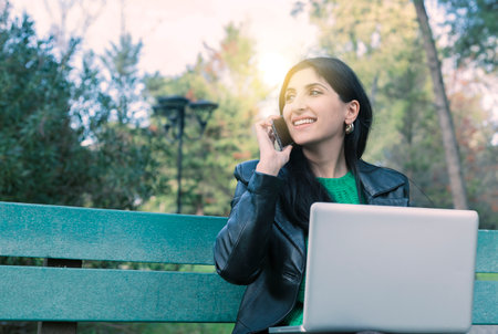 Young smiling fun student freelancer woman 20s in green jacket jeans sit on bench in spring park outdoors rest use laptop pc computer talk speak by mobile cell phone. People urban lifestyle conceptの写真素材