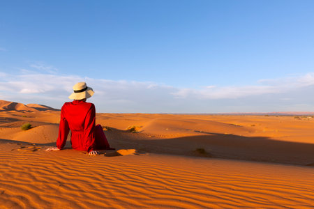 A young woman dressed in red and hat sitting on the desert dunesの写真素材
