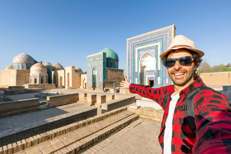 Young tourist taking a selfie at the beautiful view of the Shah-i-Zinda Ensemble in Samarkand, Uzbekistan. Mausoleums decorated with blue tiles with designs.の写真素材