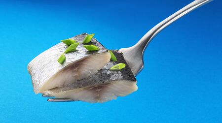 Pieces of salted herring on fork isolated on blue background, slices of marinated mackerel fish fillet with cut green onionの写真素材